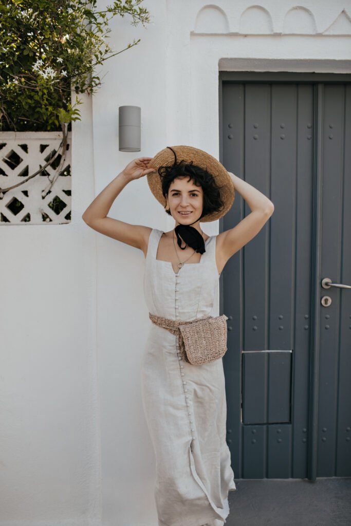 brown eyed brunette woman in white dress puts on boater outside. cheerful young lady in hat and sum